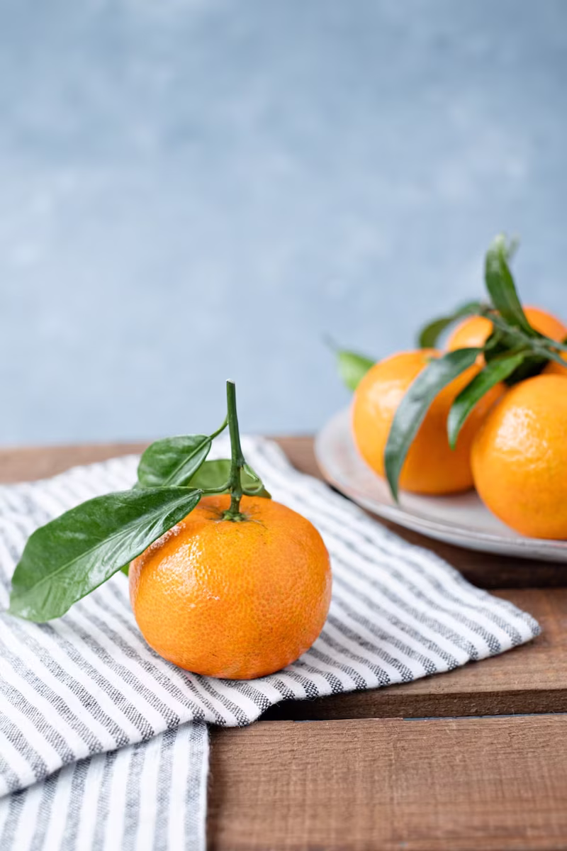 An orange with green leaves attached to them sitting atop a striped napkin. In the background there is a plate with more oranges.