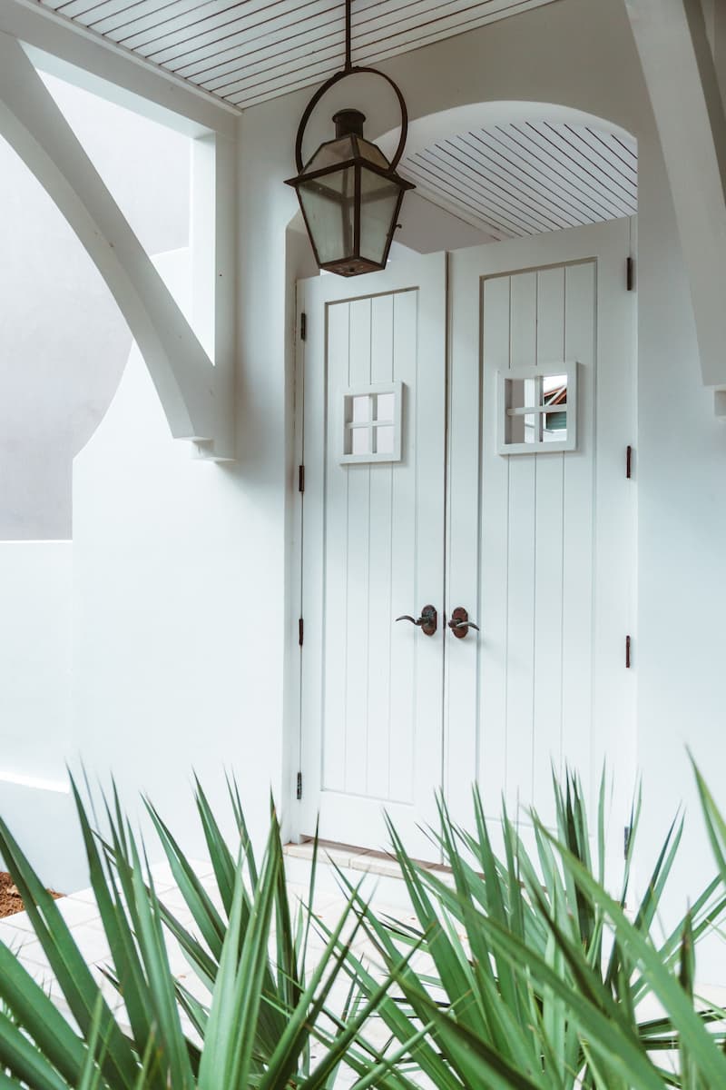 The exterior porch of a home that is all white with white front doors. A porch lantern and a small bit of greenery are also visible in the image.